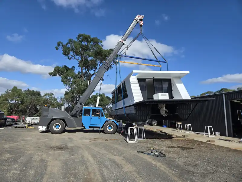 Crane with a blue cab lifting a white houseboat on a spreader frame in a gravel yard