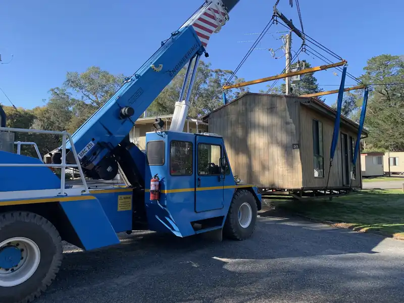 Blue Franna crane with a timber cabin slung from a spreader frame beside it
