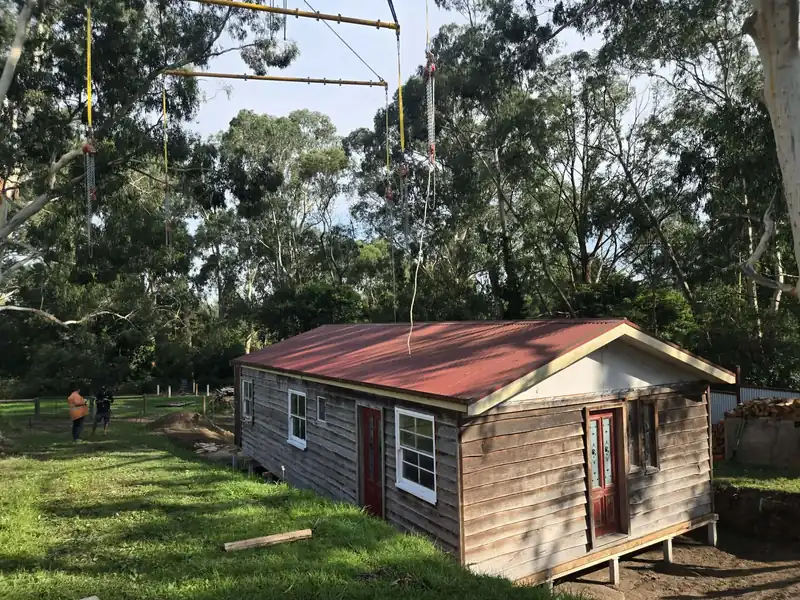 Small timber cabin with a red roof rigged to a crane for a lift on a grassy block
