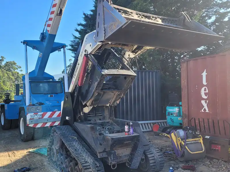 Tracked skid steer loader with its bucket raised in a yard, blue crane behind it