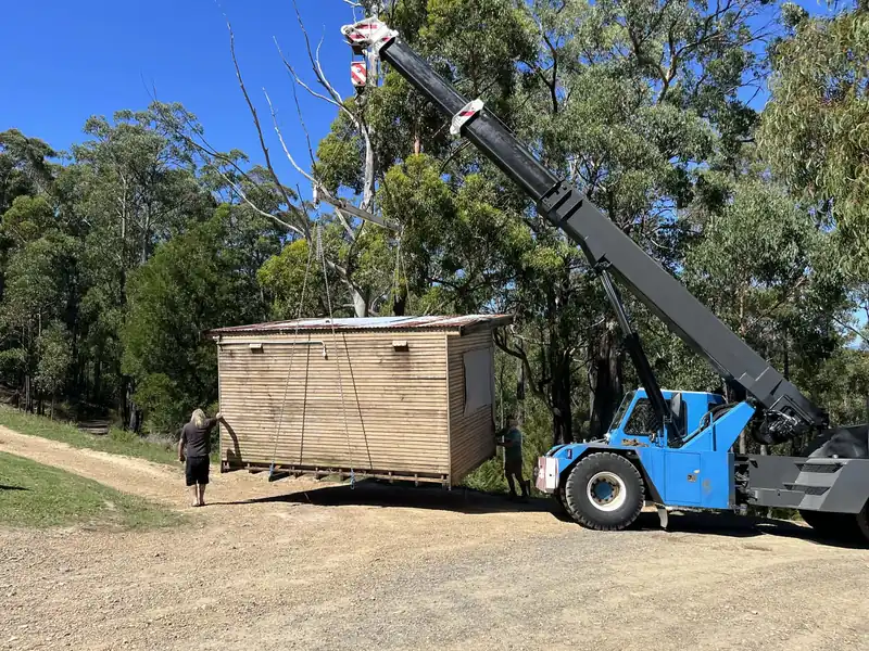 Blue Franna crane carrying a timber cabin along a gravel driveway while a worker guides it
